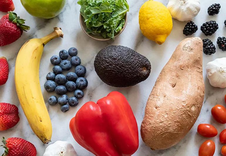 On a marble counter, there are various fruits and veggies like blueberries, strawberries, a bell pepper and a sweet potato.