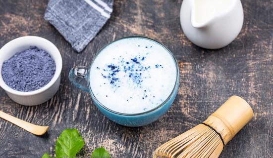 Blue matcha powder and matcha-making equipment surround a glass mug of blue matcha tea