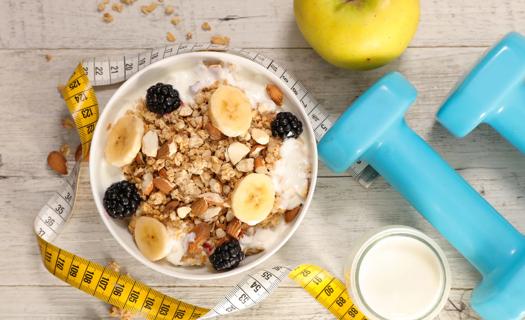 Bowl of oatmeal, bananas, blackberries and nuts, with milk, dumbbells and measuring tape on table