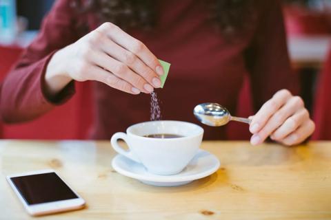 Person pouring packet of sugar subsitute in cup of coffee, cell phone on table, spoon in hand