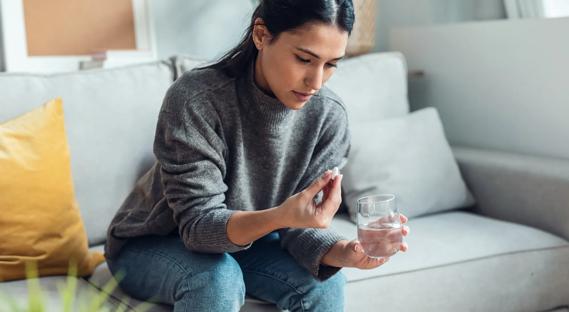 Woman sitting on couch at home, holding a pill and a glass of water, about to take the medication