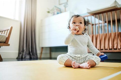 Baby sitting on floor by crib, playing with blocks, with one in their mouth